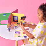 Young girl holding a dressed paper doll beside other characters and a 3D toy house set.