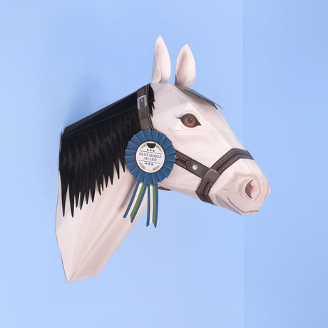 3D papercraft white horse head model mounted on a light blue wall, wearing a bridle and a blue award ribbon.