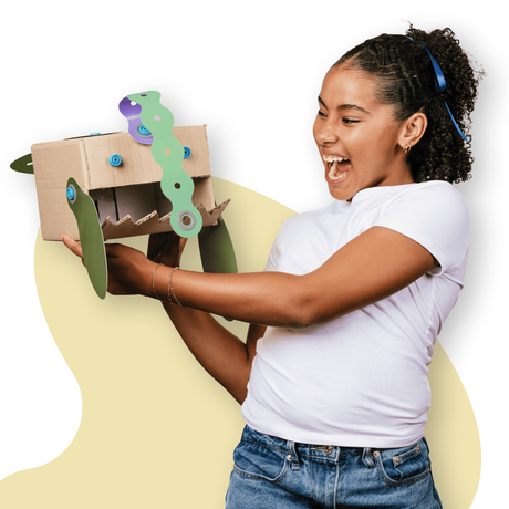 An excited girl holding a cardboard monster creation with green and purple decorative paper shapes attached.