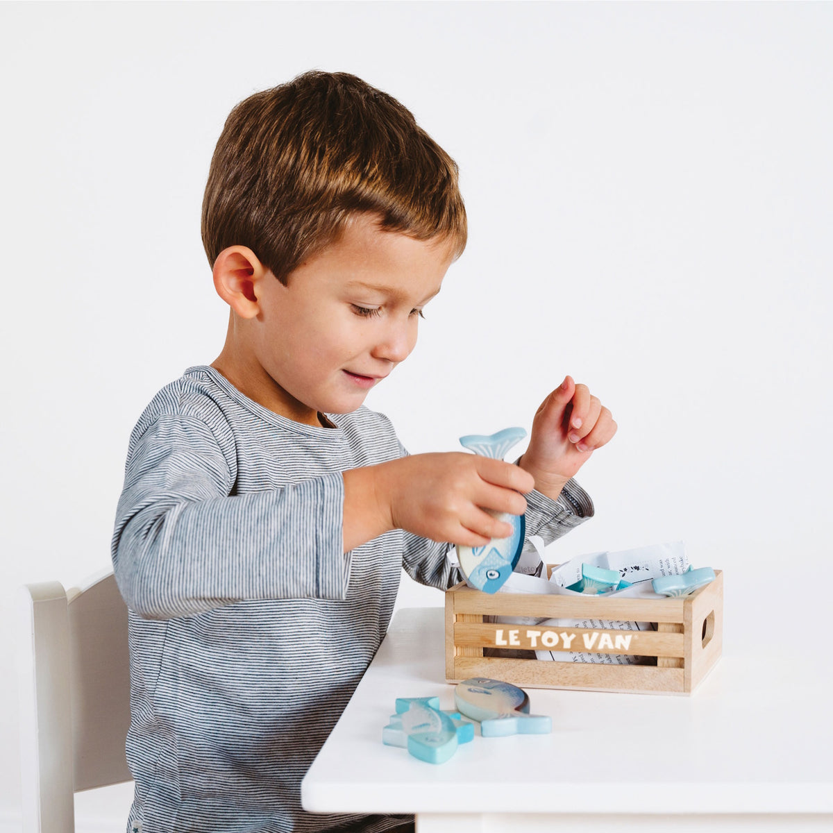 Young boy sitting at a table playing with wooden toy fish from a Le Toy Van crate.