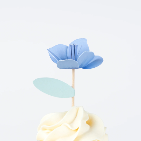 Blue flower cupcake topper with a mint green leaf on a wooden pick, resting on swirled frosting.