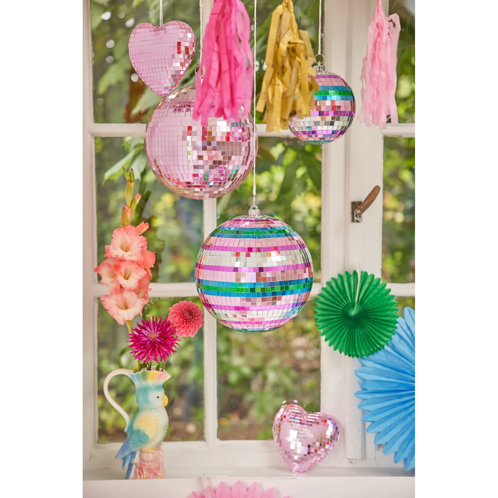 Colorful disco balls and heart ornaments hang near a window, surrounded by pink and yellow tassels. Vibrant flowers in a parrot vase add to the festive vibe.