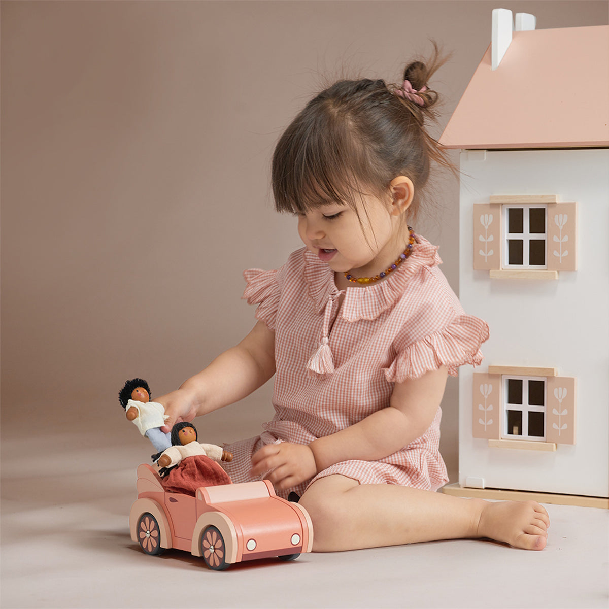 Young girl playing with wooden dolls and an orange toy car beside a dollhouse.