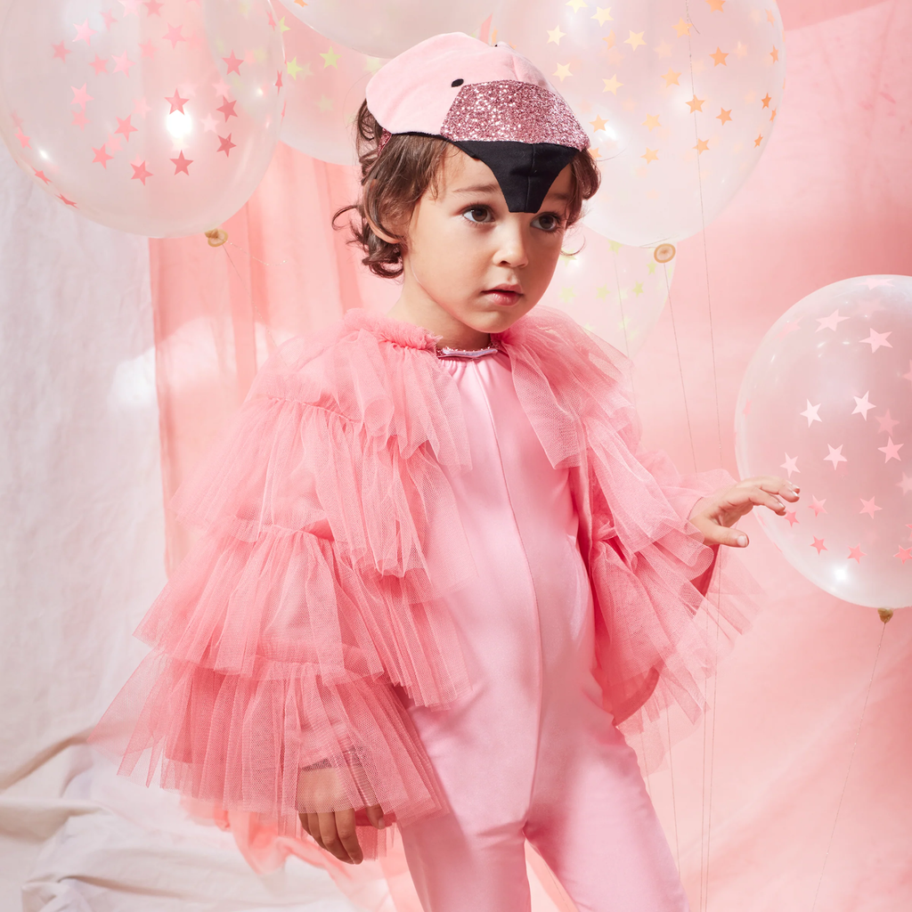 Child in a pink flamingo costume with ruffled tulle layers, wearing a matching cap. Surrounded by star-patterned balloons, set against a pink backdrop. The expression is curious.