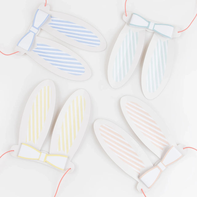Four sets of pastel striped paper bunny ears with matching bow ties and pink elastic strings on a white background.