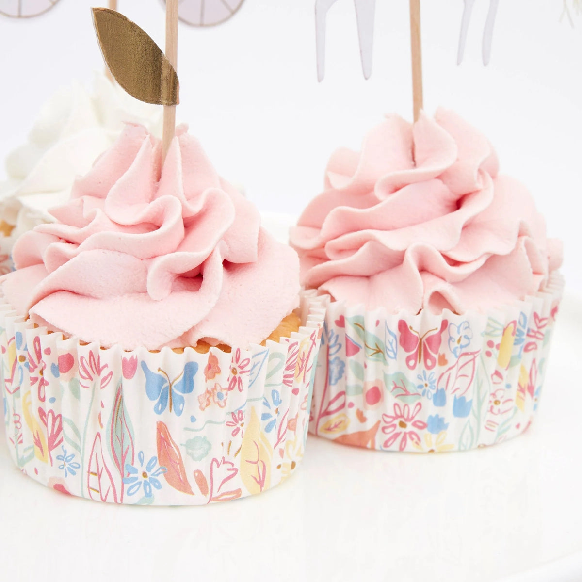 Two frosted pink cupcakes in patterned floral liners with wooden picks, on a white background.