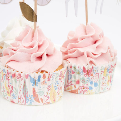 Two frosted pink cupcakes in patterned floral liners with wooden picks, on a white background.