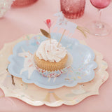 A cupcake with pink frosting, a unicorn topper, and a flower topper on a blue paper plate.