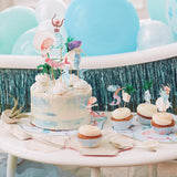 A mermaid-themed birthday cake and cupcakes decorated with toppers against a blue balloon background.