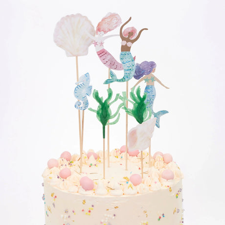 A frosted cake topped with mermaid, seashell, seahorse, and seaweed paper toppers on sticks.