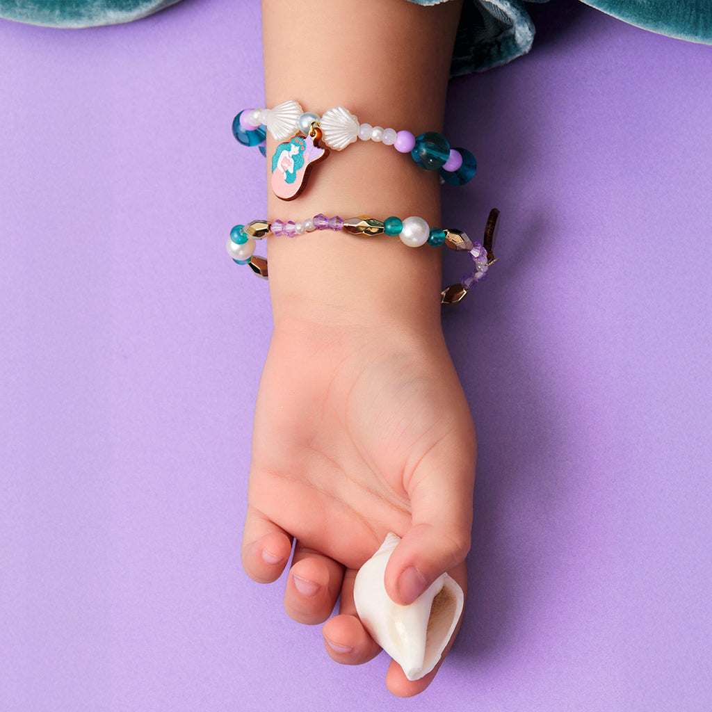 A child's hand rests on a purple surface, wearing colorful bracelets with beads and charms, holding a small white seashell. The mood is playful and gentle.