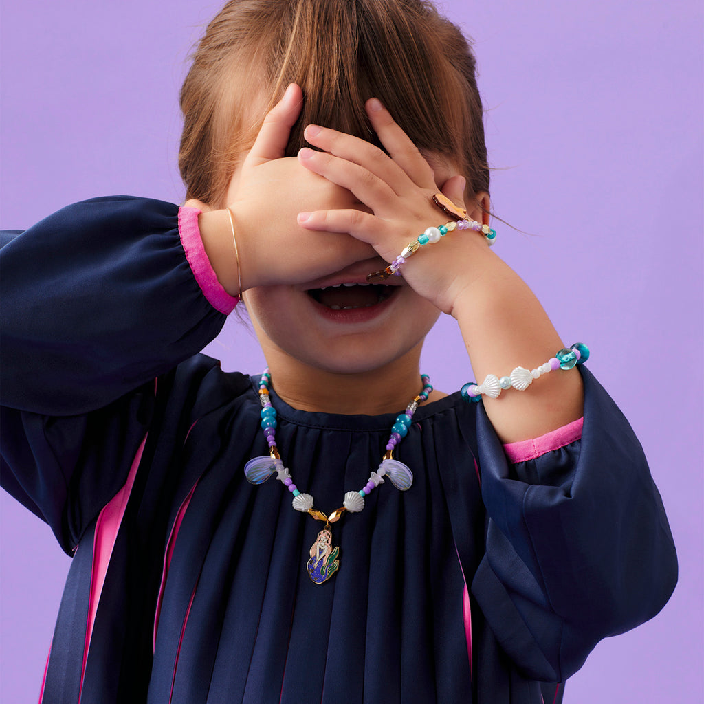 A child in a navy dress with pink accents playfully covers their face with their hands. They wear colorful bead jewelry against a pastel purple background.