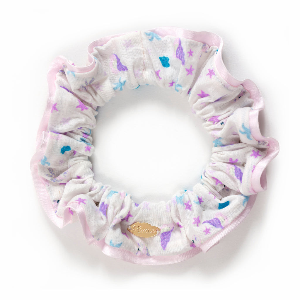 A pastel-colored scrunchie with ruffled edges, featuring purple and blue aquatic patterns on white fabric and a small gold tag, on a white background.