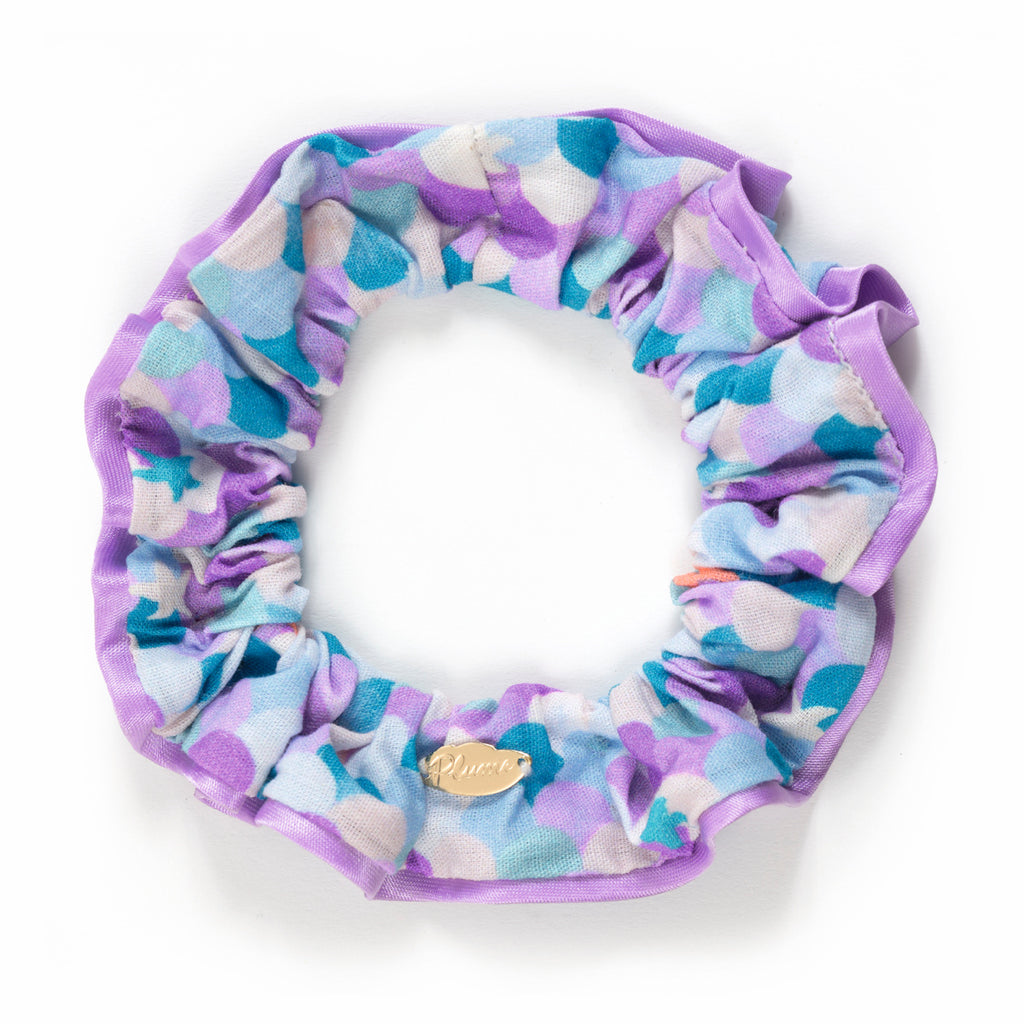 A colorful scrunchie with a pattern of purple, blue, and white circles. It has a gold tag attached. The design is vibrant and playful.