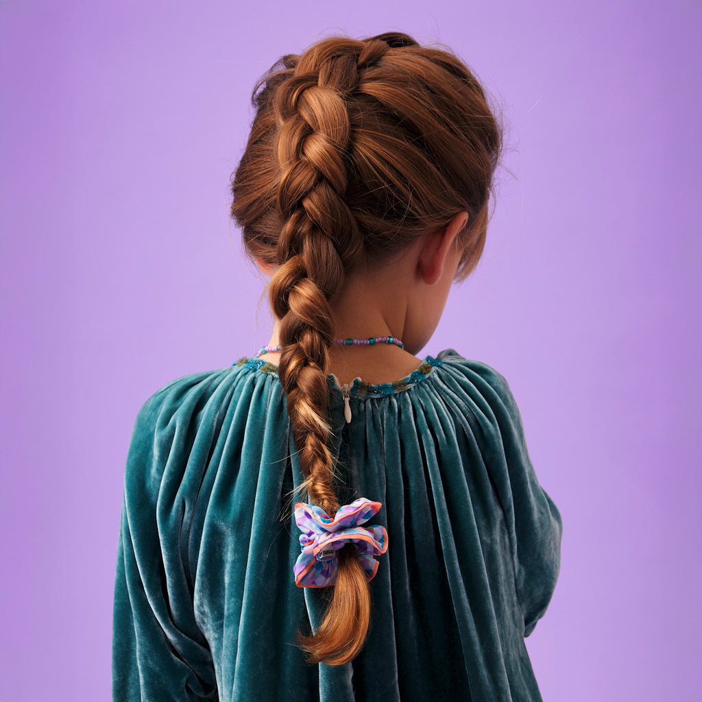 Young girl with a meticulously braided brown hair, tied with a colorful scrunchie. She wears a teal velvet dress, set against a soft purple backdrop.