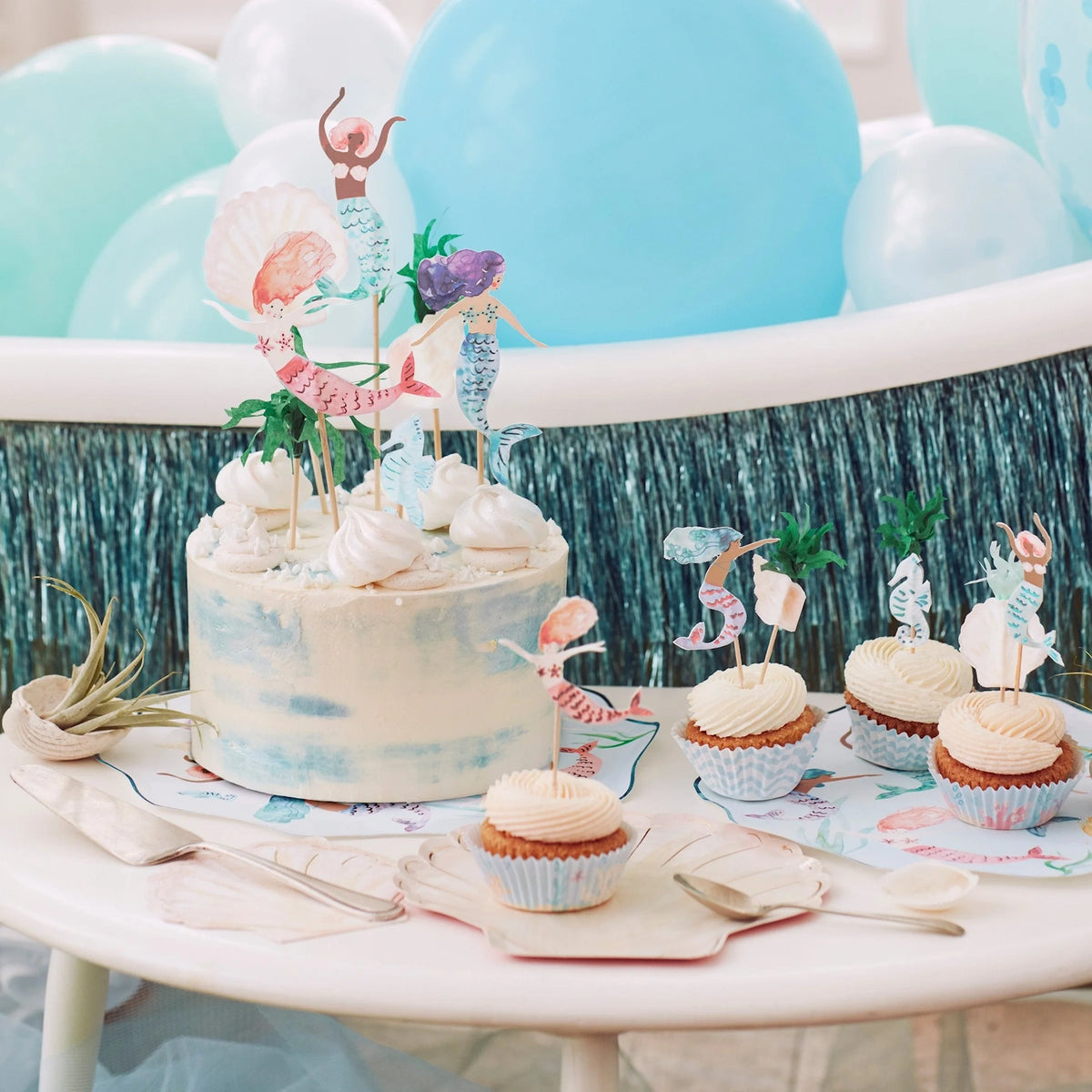 A mermaid-themed birthday cake and cupcakes on a table decorated with blue balloons and tinsel.