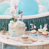 A mermaid-themed birthday cake and cupcakes on a table decorated with blue balloons and tinsel.
