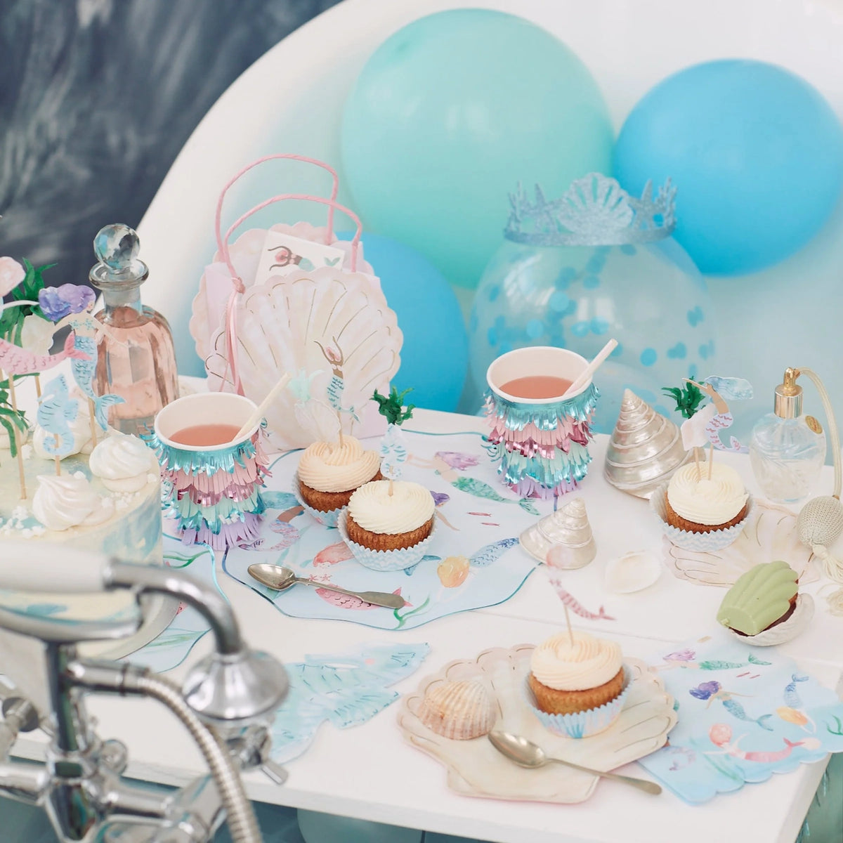 A mermaid-themed party setup with cupcakes, fringed cups, and blue balloons by a bathtub.