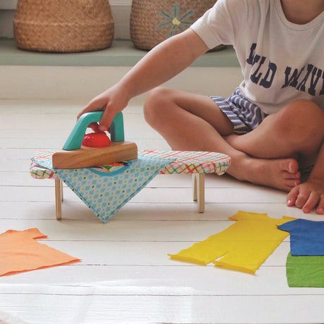 Child using a wooden toy iron and ironing board set on a white floor with colorful fabric clothes.