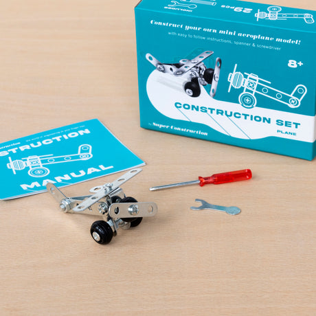 A construction set with a partially assembled toy plane is on a table. Nearby are a blue instruction manual, a red screwdriver, and a small wrench.