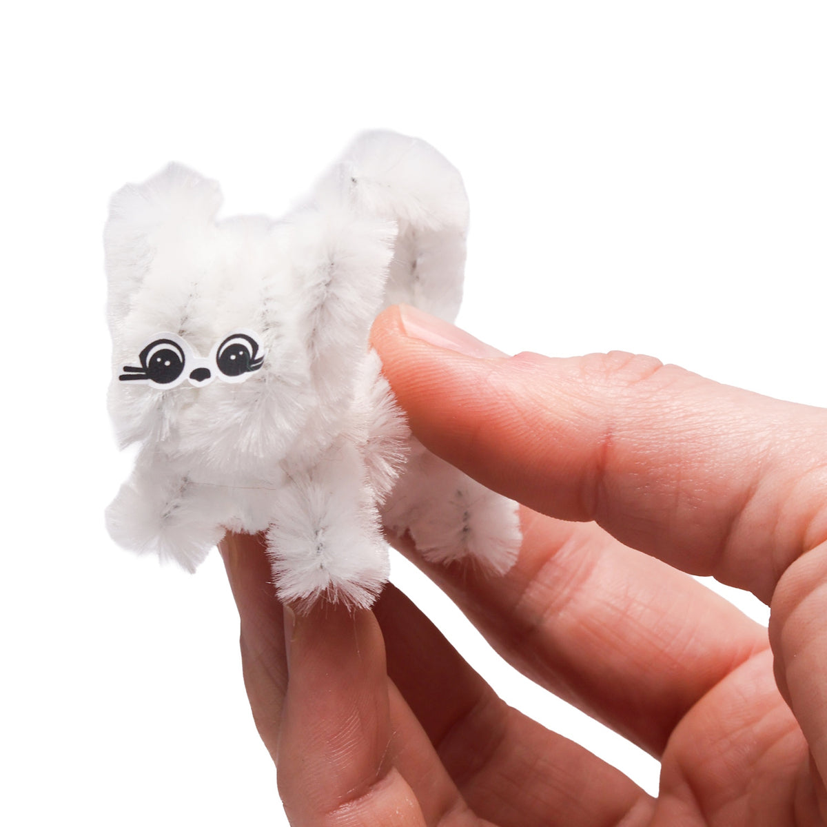 Hand holding a tiny white fluffy pipe cleaner cat figure with paper eye stickers.