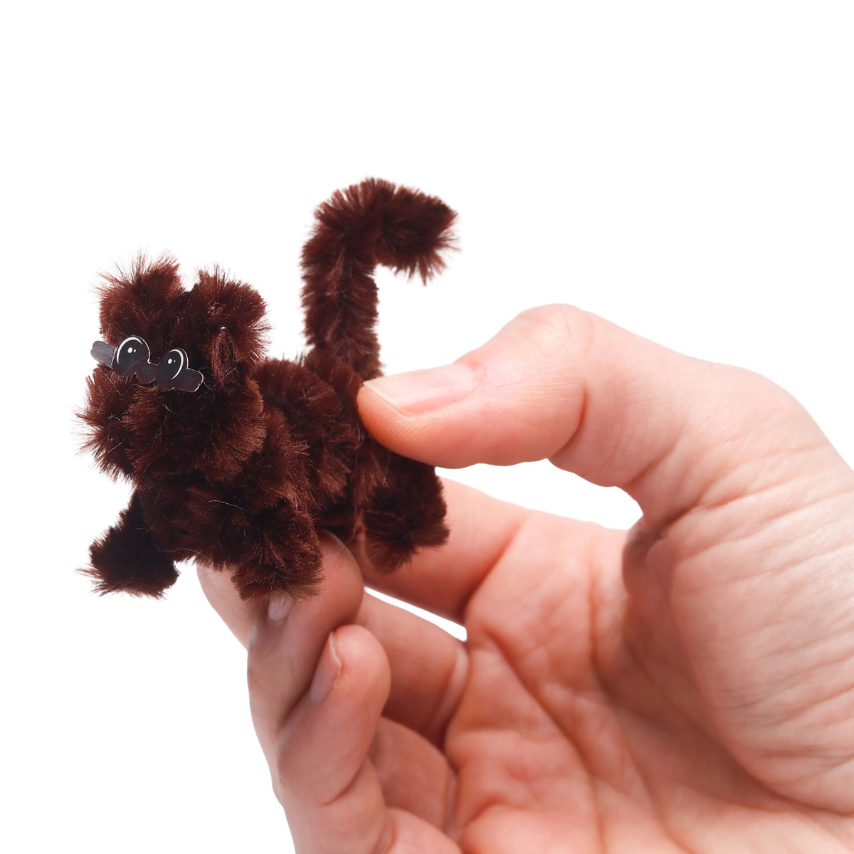 Hand holding a small, brown fuzzy pipe cleaner animal with sticker eyes on a white background.