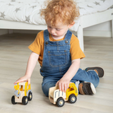 A child with curly hair, wearing blue overalls and a yellow shirt, sits on a wooden floor playing with two yellow toy trucks, focused and content.
