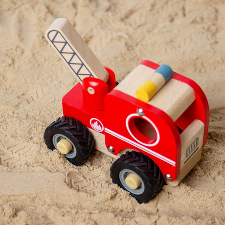 Wooden toy fire truck in bright red with black wheels on sandy ground. The playful setting evokes a cheerful and imaginative tone.