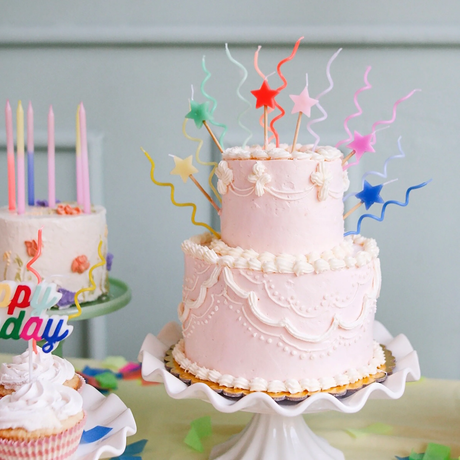 Two-tiered pink cake on a stand decorated with colorful star candles and wavy candle wicks.