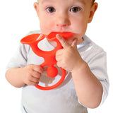 A baby in a light shirt holds a red trumpet-shaped teething toy, gently biting it. The child appears curious and focused, set against a plain background.