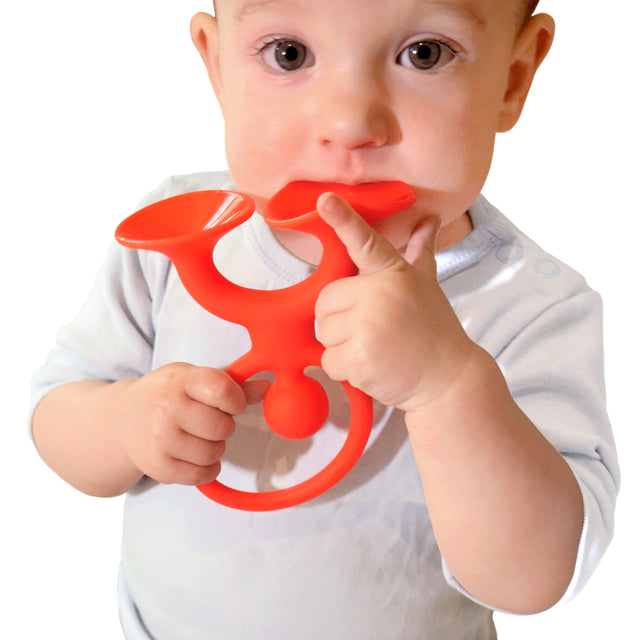 A baby in a light shirt holds a red trumpet-shaped teething toy, gently biting it. The child appears curious and focused, set against a plain background.