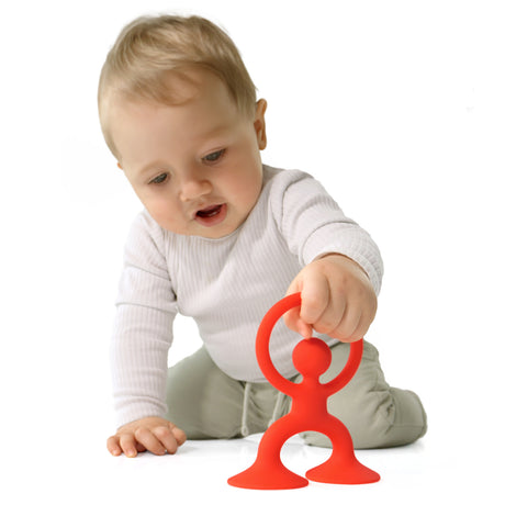 A curious baby in a white shirt and light green pants plays with an orange toy resembling a human figure on a white background, evoking a sense of exploration.