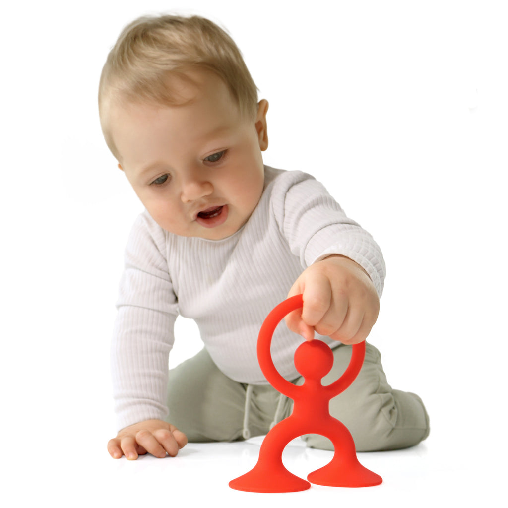 A curious baby in a white shirt and light green pants plays with an orange toy resembling a human figure on a white background, evoking a sense of exploration.