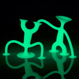 Two green, glowing abstract figures with round bases pose playfully against a black background, casting reflections on a shiny surface.