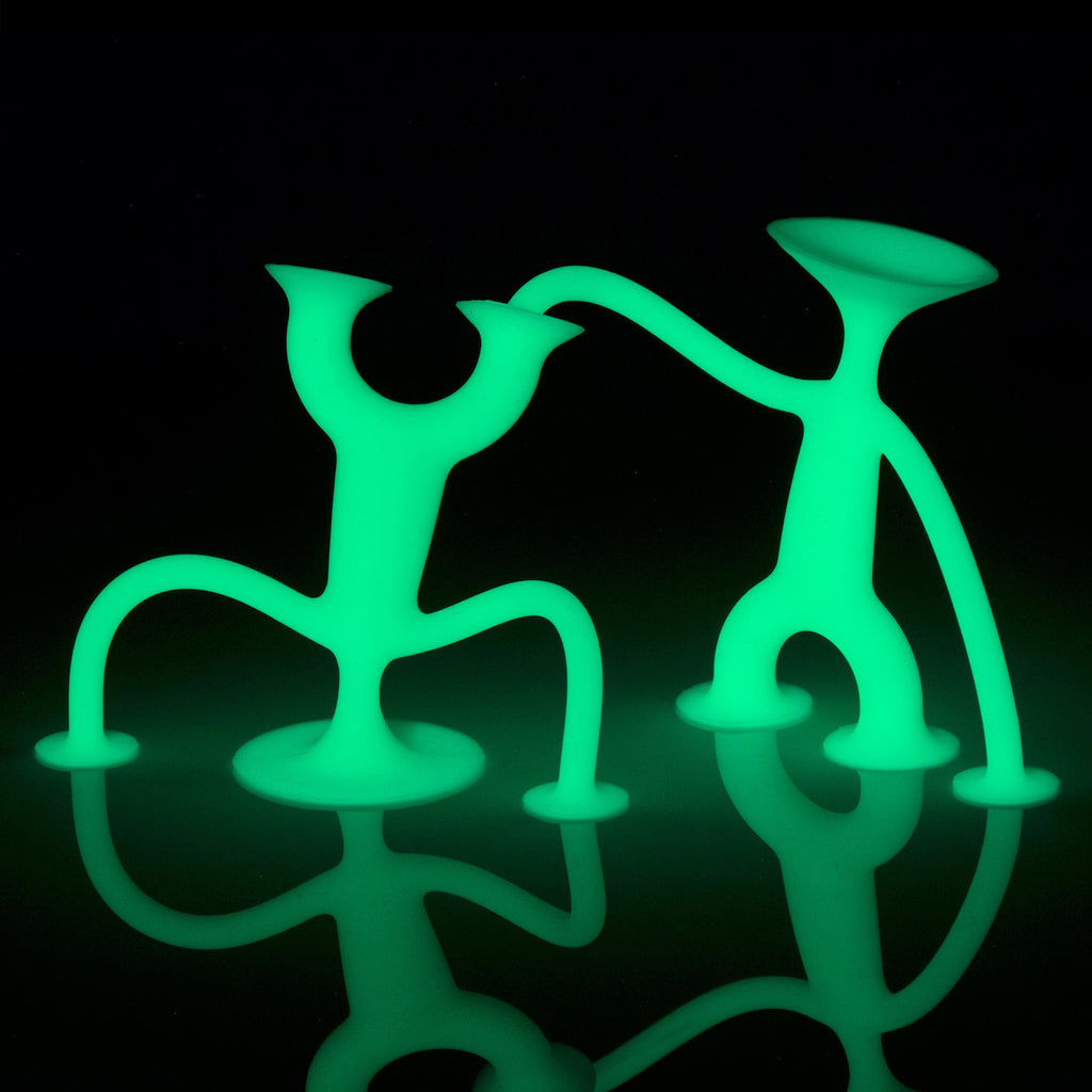 Two green, glowing abstract figures with round bases pose playfully against a black background, casting reflections on a shiny surface.