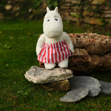 A plush toy resembling a hippo stands on rocks in a garden, wearing a red and white striped skirt. The background features a stone wall and greenery.
