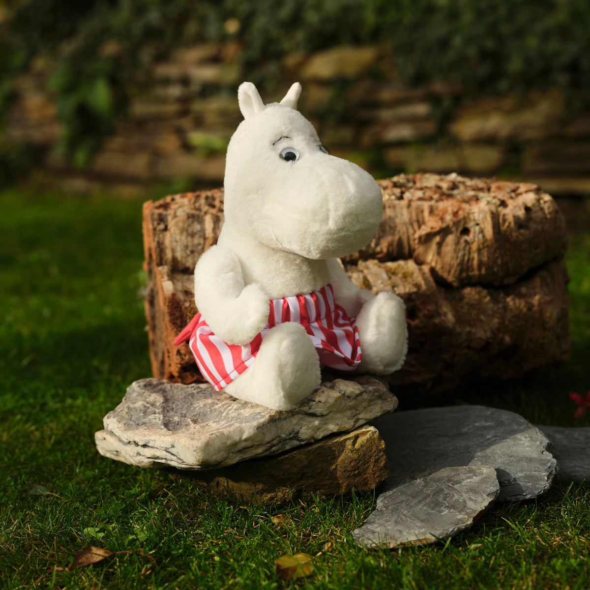 A white plush toy resembling a hippopotamus, wearing red and white striped shorts, sits on a stone in a garden with green grass and a brick wall.