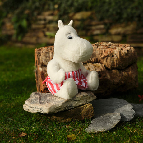 A white plush toy resembling a hippopotamus, wearing red and white striped shorts, sits on a stone in a garden with green grass and a brick wall.