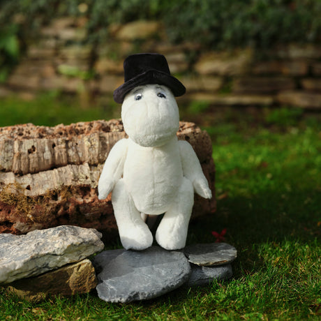 A white plush toy with a black hat sits on gray stones in front of a textured log. The scene is set on a grassy surface, conveying a whimsical tone.
