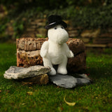 A white plush toy wearing a black hat sits on stones in a garden, backed by a rustic wooden log. The scene exudes a whimsical and peaceful vibe.