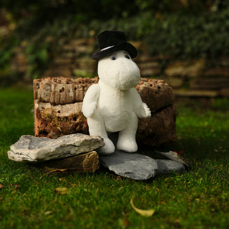 A white plush toy wearing a black hat sits on stones in a garden, backed by a rustic wooden log. The scene exudes a whimsical and peaceful vibe.