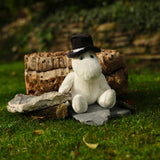 A plush toy wearing a black top hat sits on a stone slab in a grassy garden. Behind it, there's a log and foliage, creating a whimsical outdoor scene.