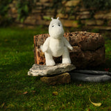 A white plush toy with a friendly expression sits on stone slabs in a garden, with lush green grass and a rustic stone wall in the background.