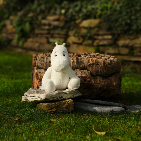 A white plush hippo sits on stacked stones in a lush green garden, with a rustic stone wall and foliage in the background, creating a peaceful, whimsical ambiance.