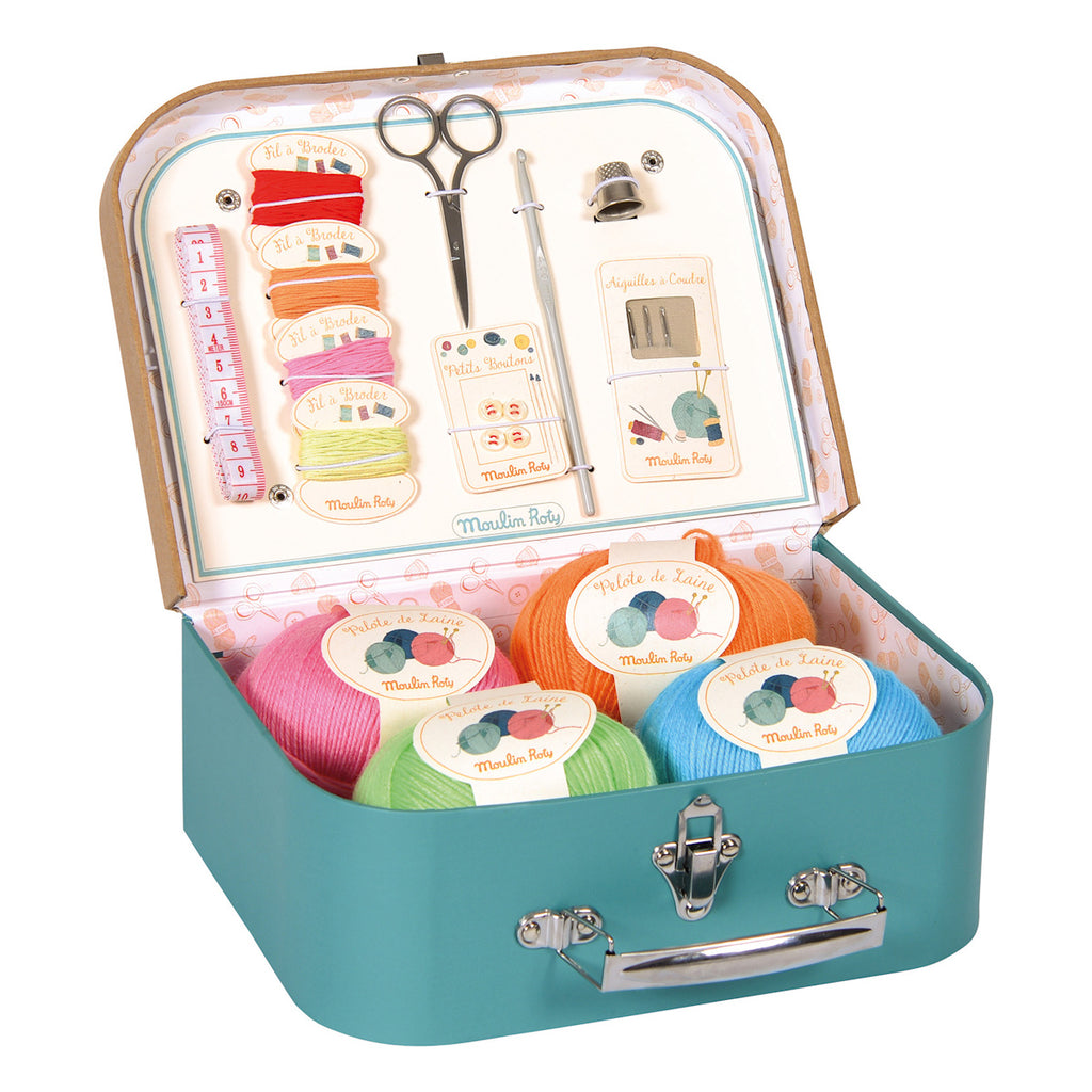 A teal sewing kit suitcase contains colorful yarn balls, scissors, tape measure, needles, and a crafting guide. The scene is creative and organized.