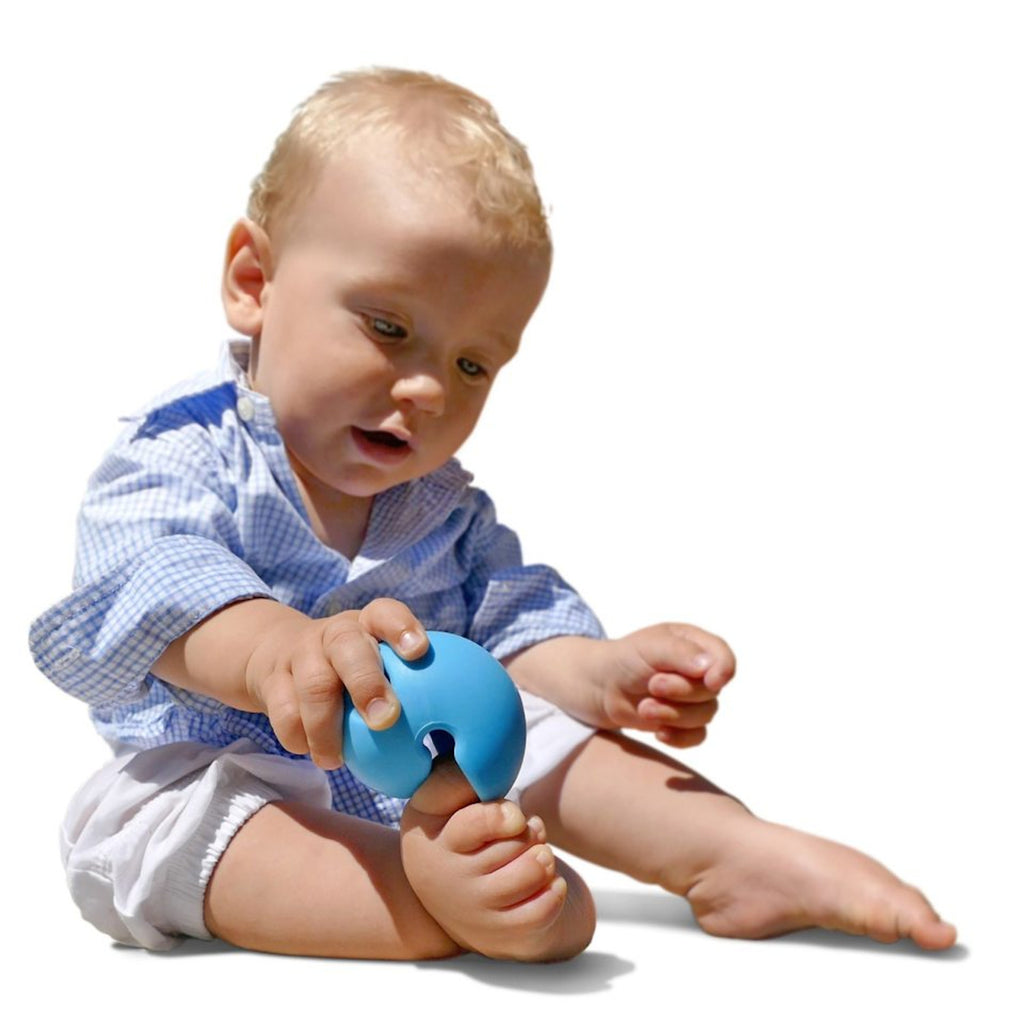 A baby in a blue checkered shirt and white shorts sits on the floor, holding a blue teething toy. The expression is curious and engaged.