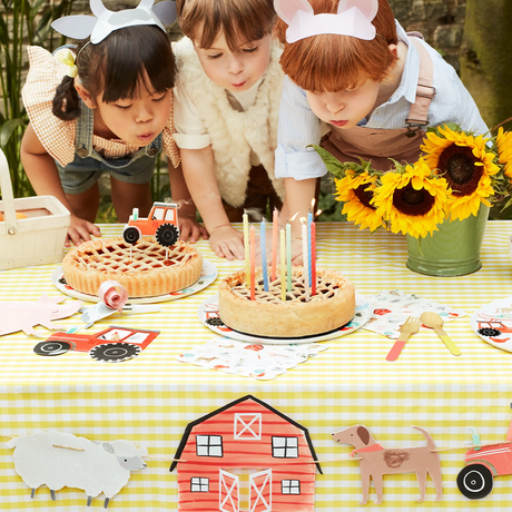 Three children blowing out candles on a pie at a farm-themed birthday celebration outdoors.