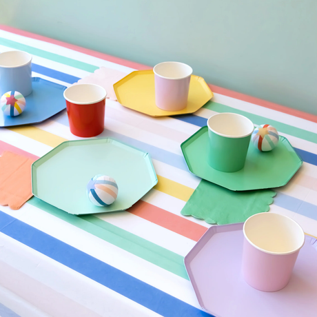 A colorful party table setup featuring octagonal paper plates, matching cups, napkins, and surprise balls arranged on a pastel striped tablecloth.