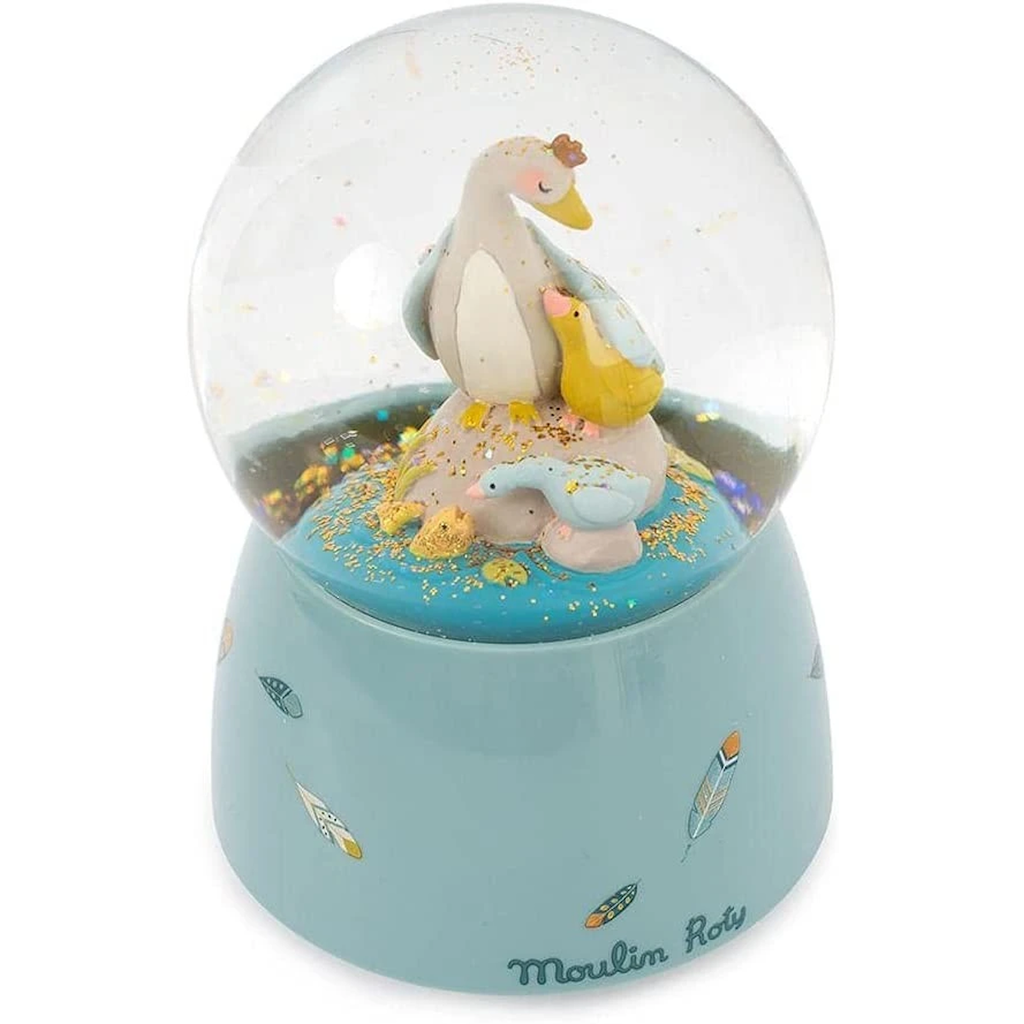 A snow globe containing a pastel-colored duck family stands on a light blue base adorned with colorful leaf illustrations. The brand name "Moulin Roty" is visible.