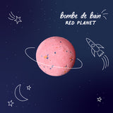 Pink bath bomb resembling a planet with doodles of a crescent moon, stars, and a rocket on a dark blue background. Text reads "bombe de bain, Red Planet."